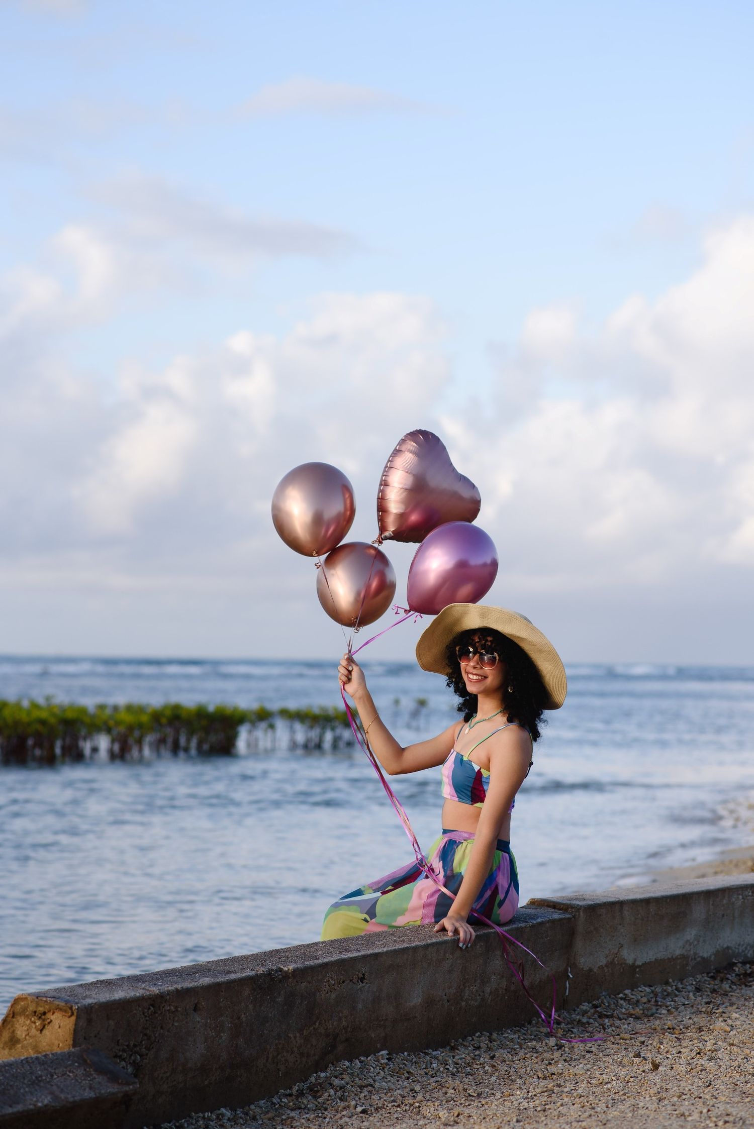 Woman in straw hat holding pink heart balloons sitting on beach wall with ocean backdrop.