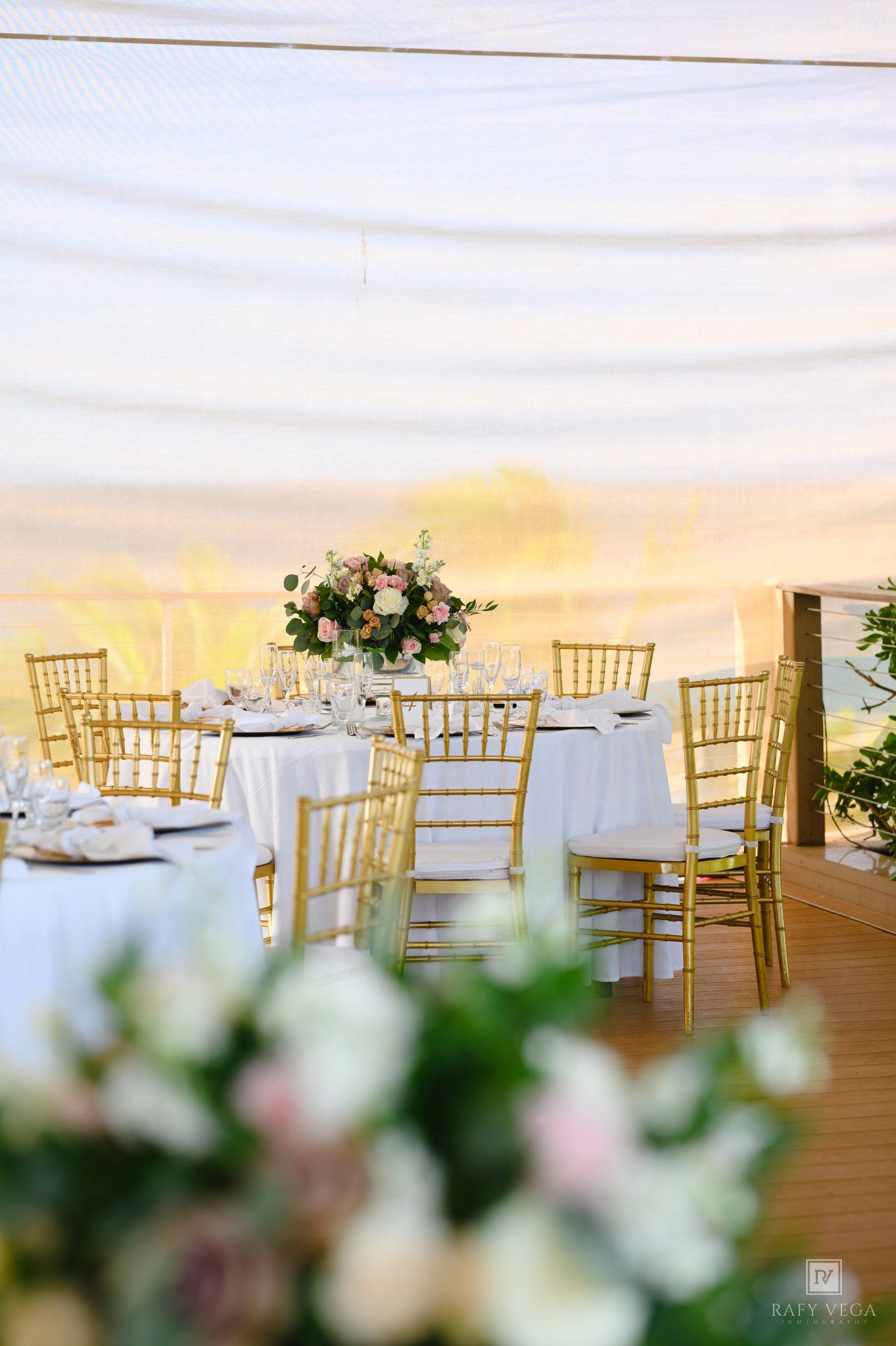 Elegant outdoor wedding reception with gold chiavari chairs, white tablecloths, and pink floral centerpieces under a draped tent.