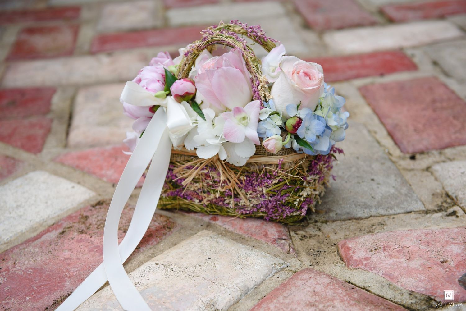 A rustic wicker basket adorned with pink roses, hydrangeas, and wildflowers with a white ribbon on brick pavers.