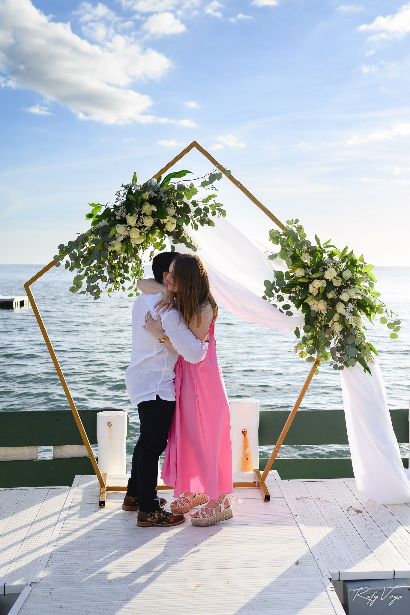 Couple kissing under a gold geometric arch with white flowers and greenery on a waterfront dock.