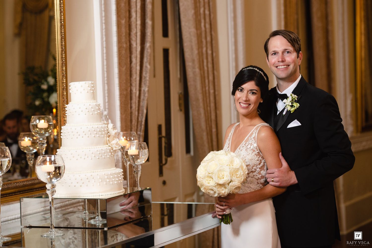 Bride and groom smiling beside a white tiered wedding cake in an elegant ballroom reception venue.