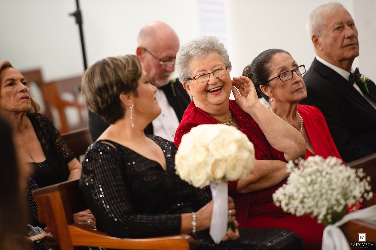 Elderly woman laughing with guests holding white bouquet at wedding ceremony.