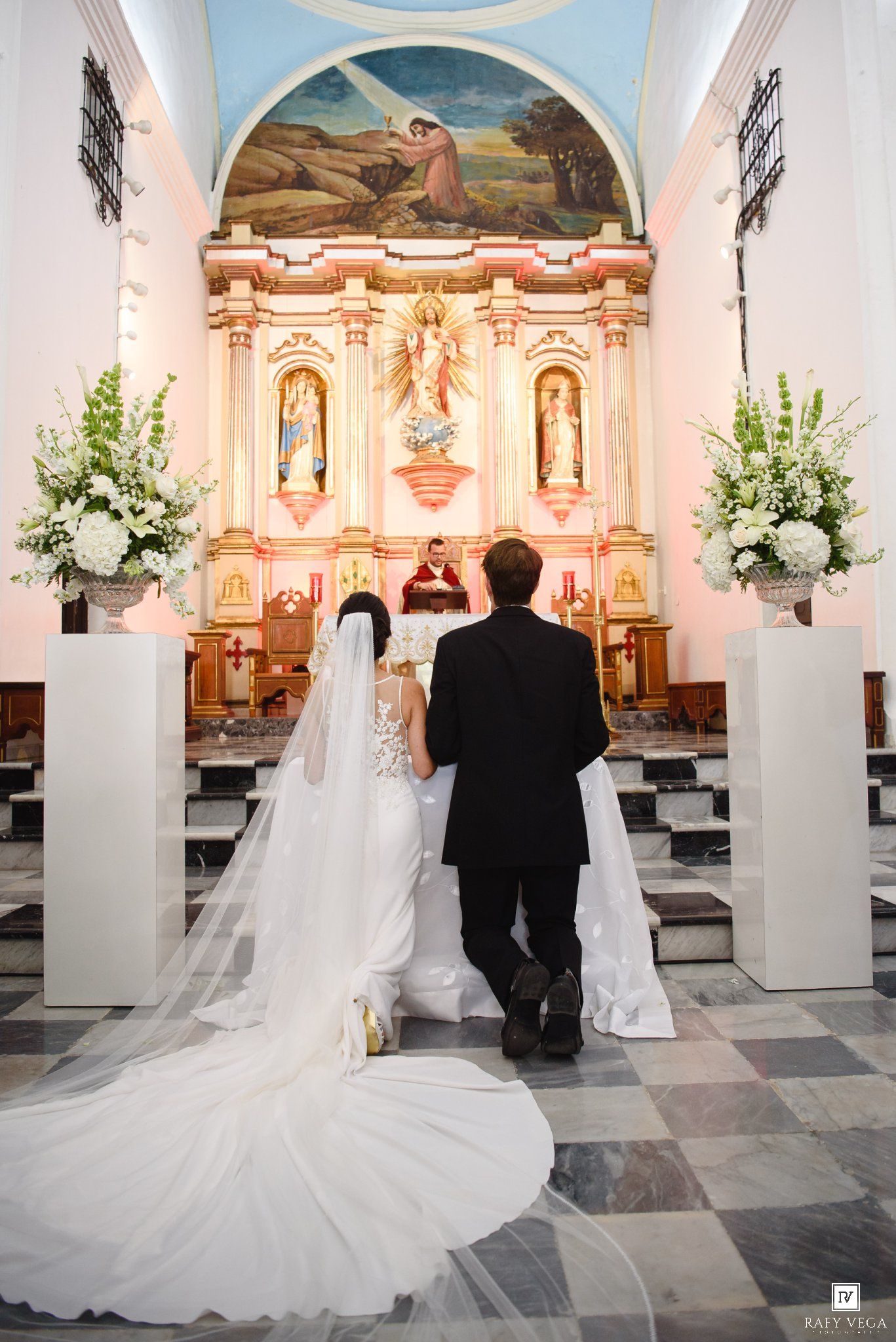 Bride and groom kneeling at ornate church altar during Catholic wedding ceremony with white floral arrangements.