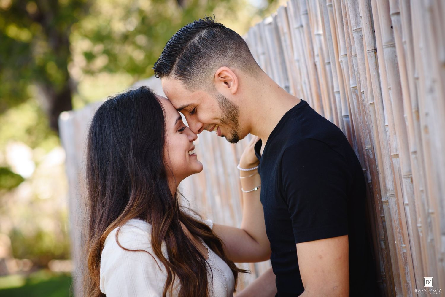 Romantic couple sharing an intimate moment, foreheads touching, smiling near a wooden fence outdoors.
