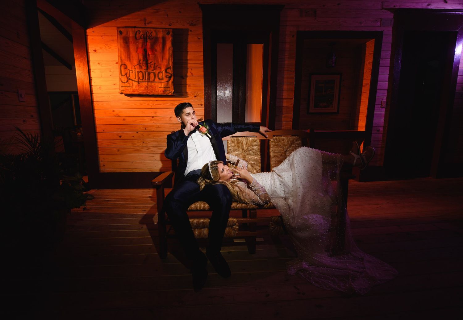 Bride and groom relax on rustic chair on wooden porch in moody purple-lit wedding venue at night.