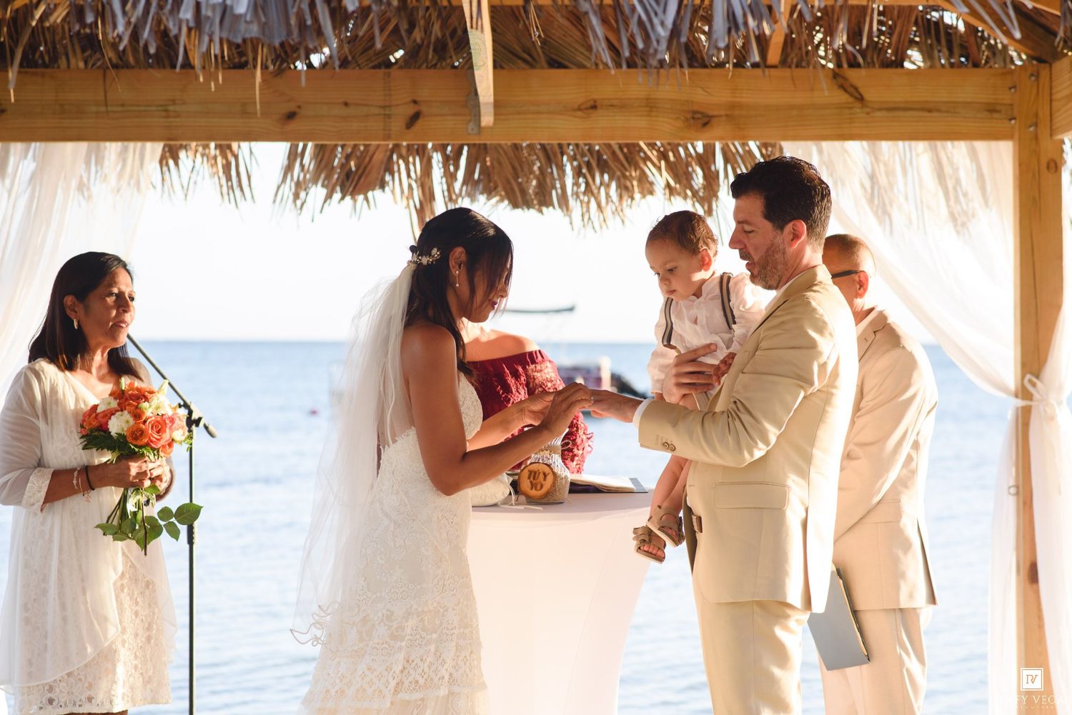 Couple exchanges vows at beachside wedding ceremony under thatched canopy, groom holding baby, officiant and guest present.