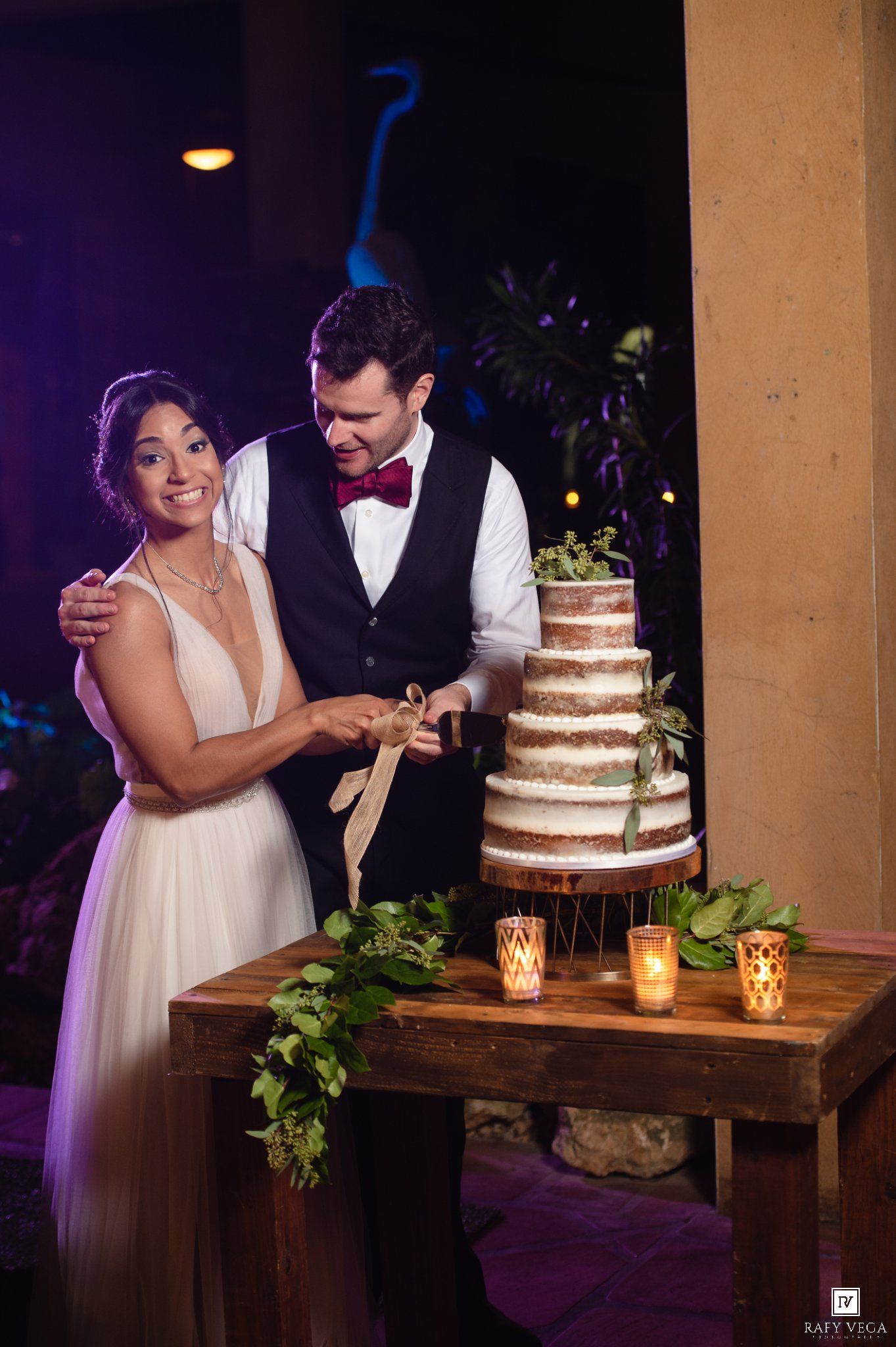 Bride and groom cutting a rustic naked wedding cake decorated with greenery, surrounded by candlelight at their reception.