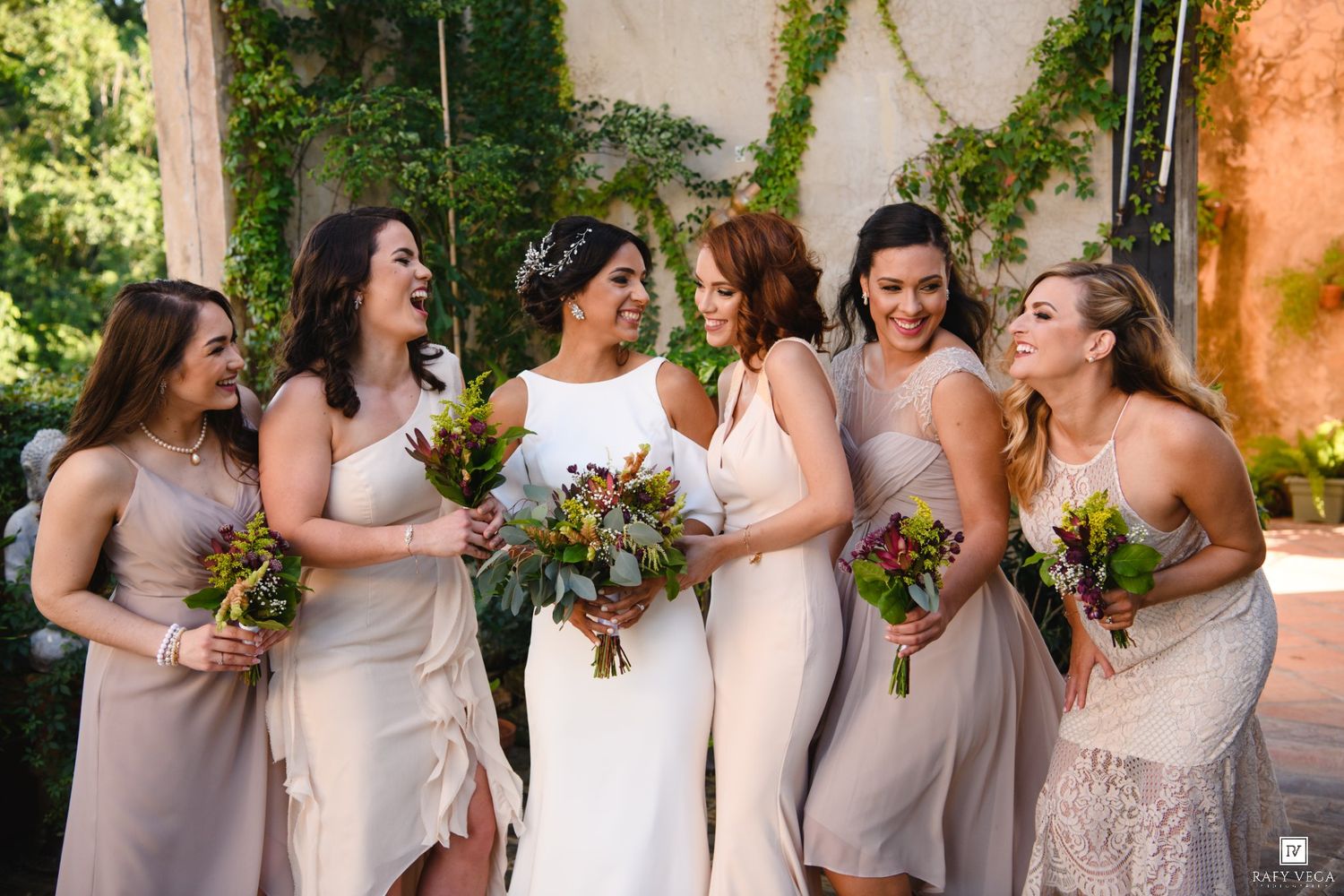 Bride in white gown laughing with five bridesmaids in champagne dresses holding colorful bouquets outdoors.