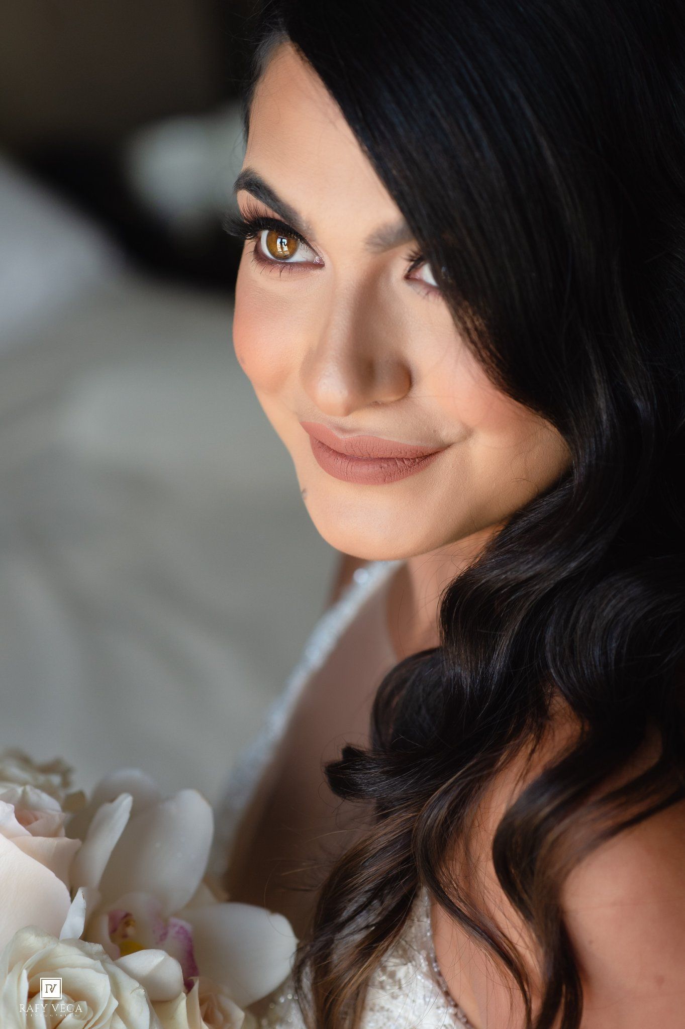 Close-up portrait of a beautiful bride with dark wavy hair, smoky eye makeup, holding white peony flowers.