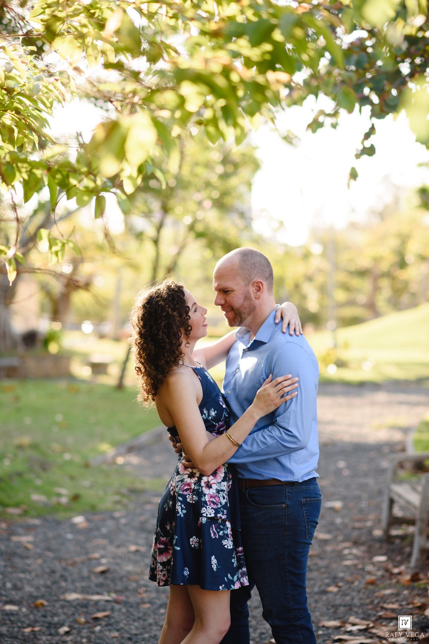 Couple embracing and gazing at each other under lush green trees in a sunlit park during an engagement photo session.