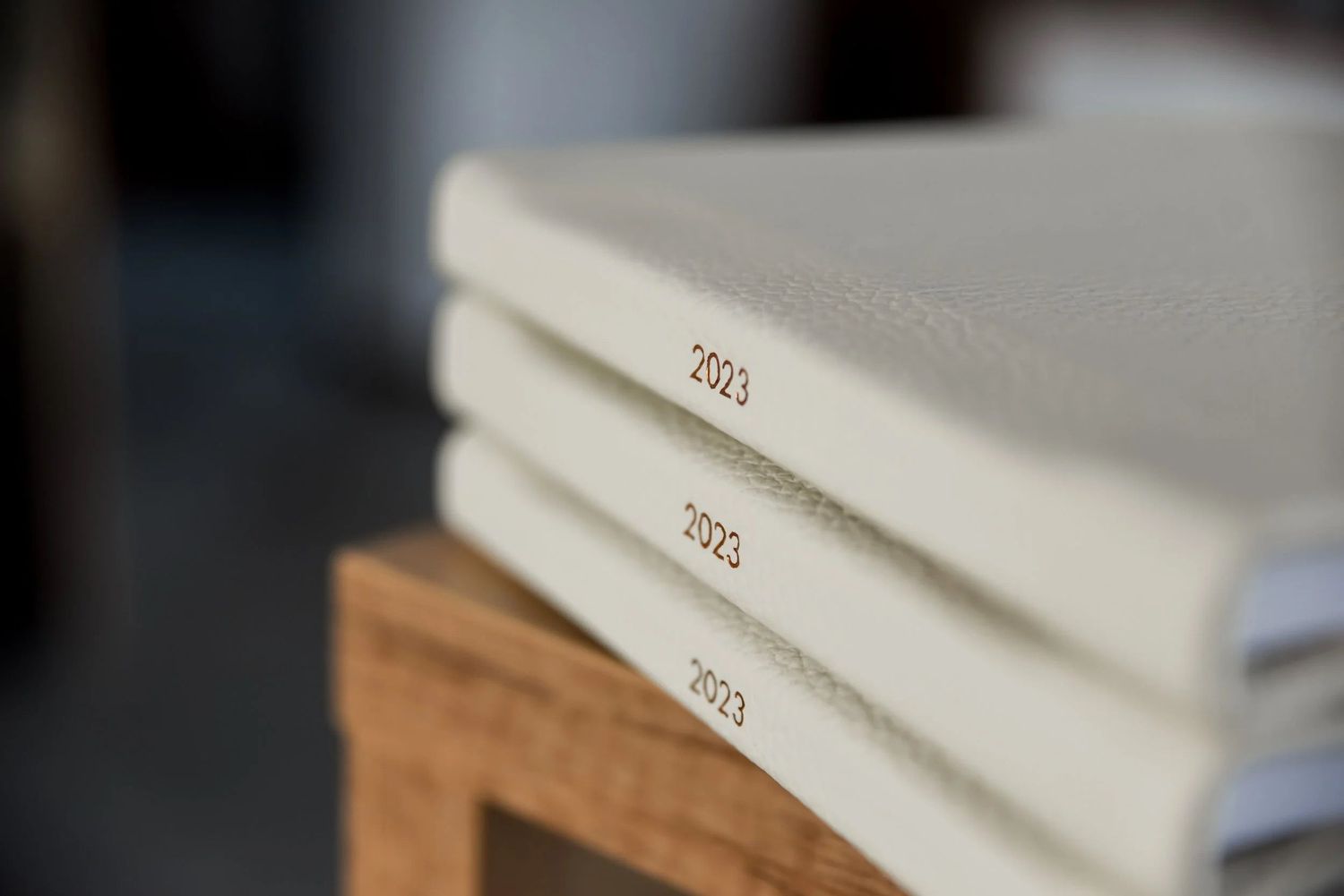 Three white 2023 hardcover journals stacked on a wooden stool with elegant gold embossed text on spines.