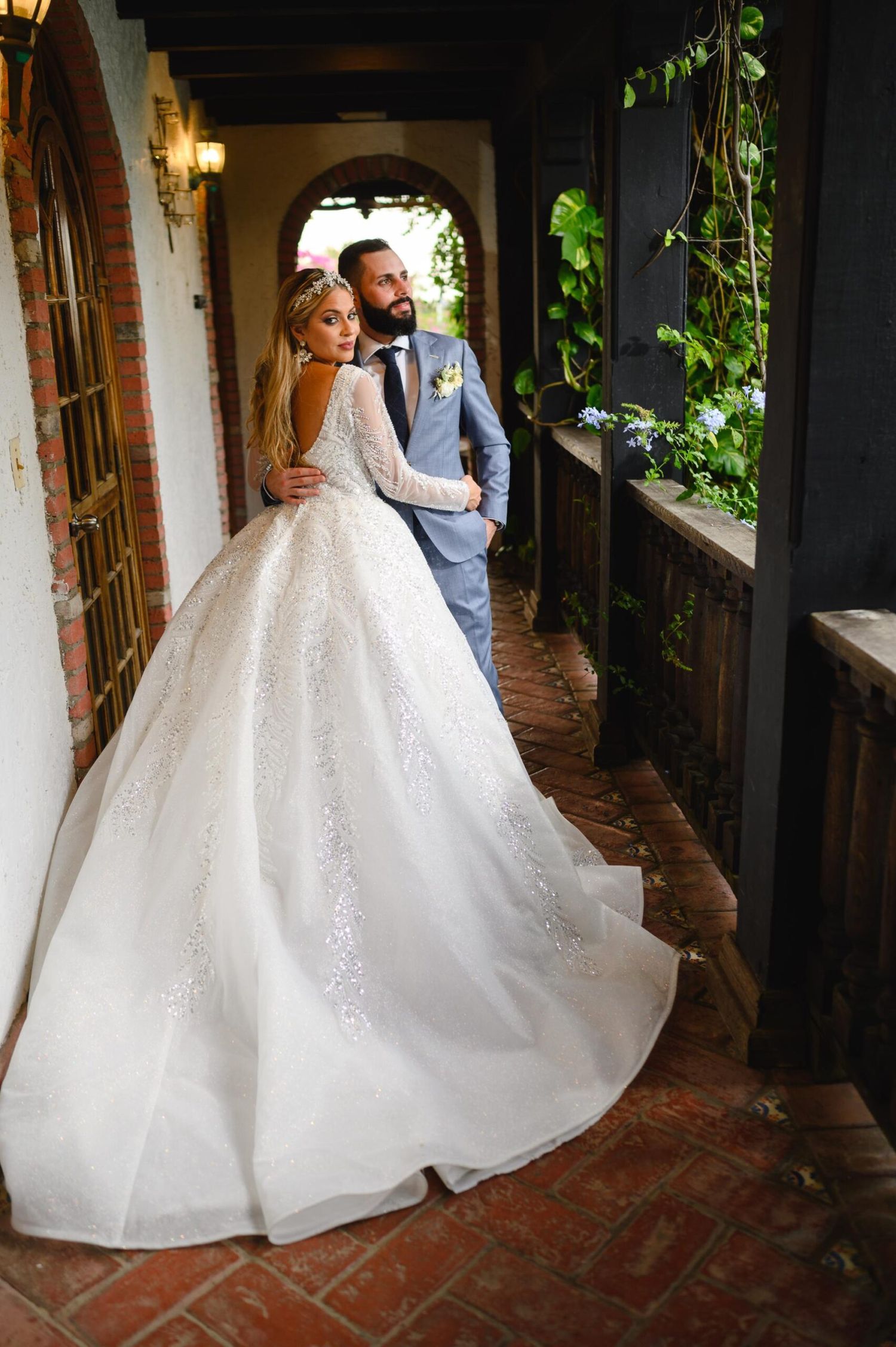 Bride in a stunning backless ball gown with long sleeves and groom in a blue suit posing in an elegant venue hallway.
