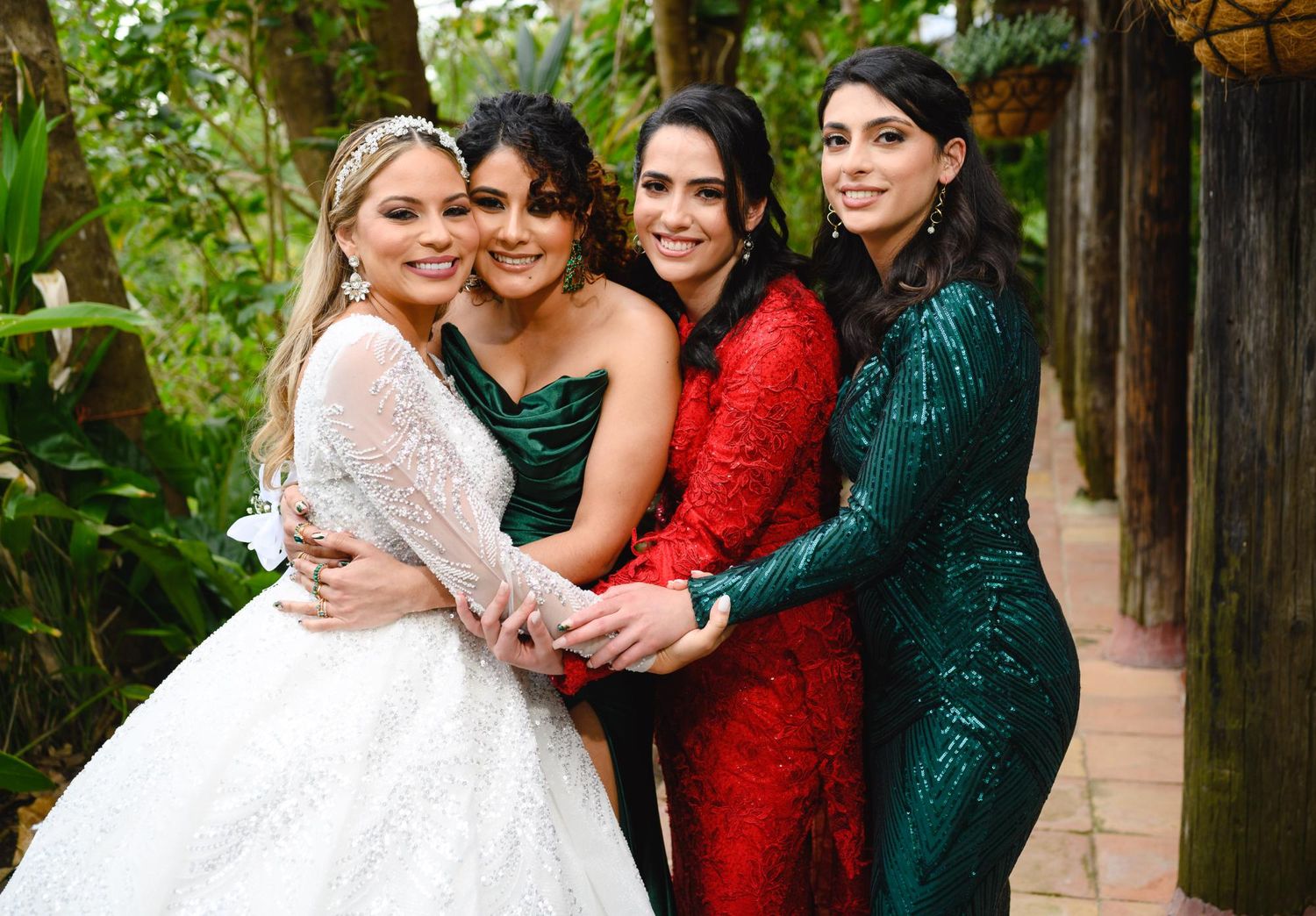 Four women in formal gowns pose together outdoors, one in a white wedding dress, others in red and green sequined dresses.