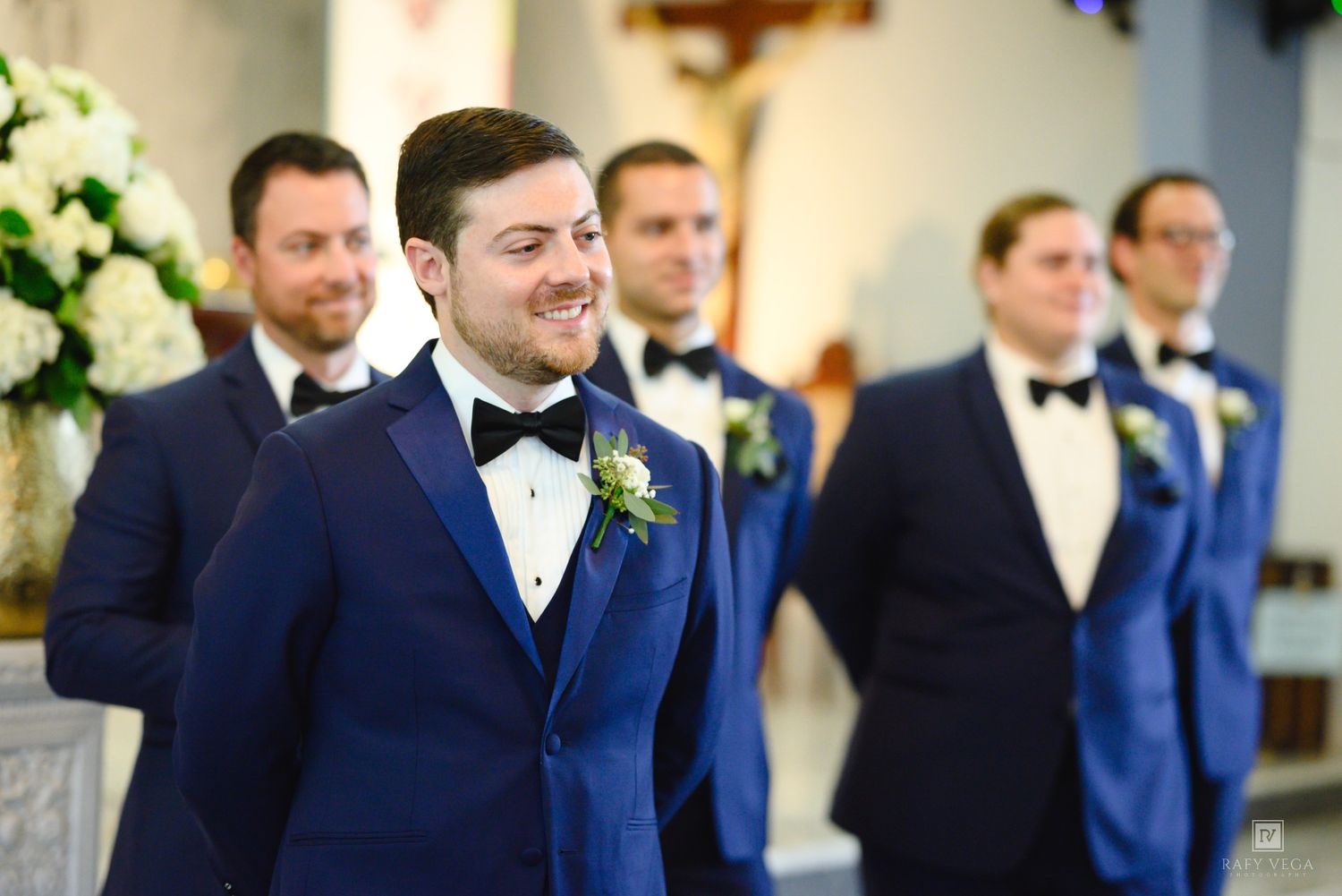 Groom in navy blue tuxedo smiling at altar with groomsmen in matching suits during church wedding ceremony.