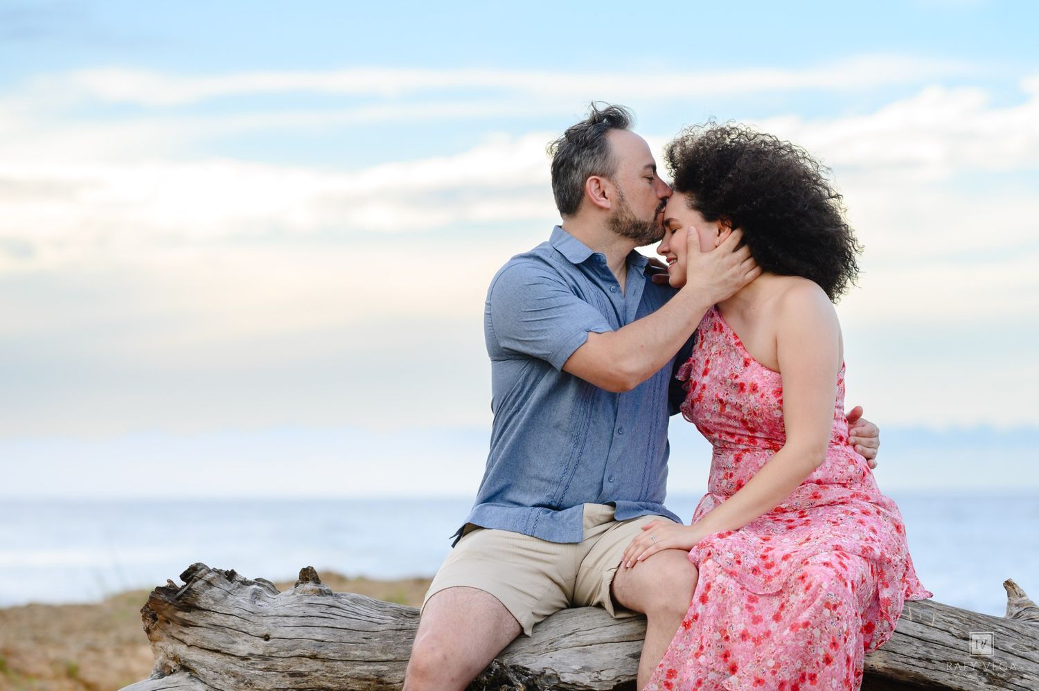 Couple sharing a romantic kiss while sitting on driftwood at the beach with ocean in the background.