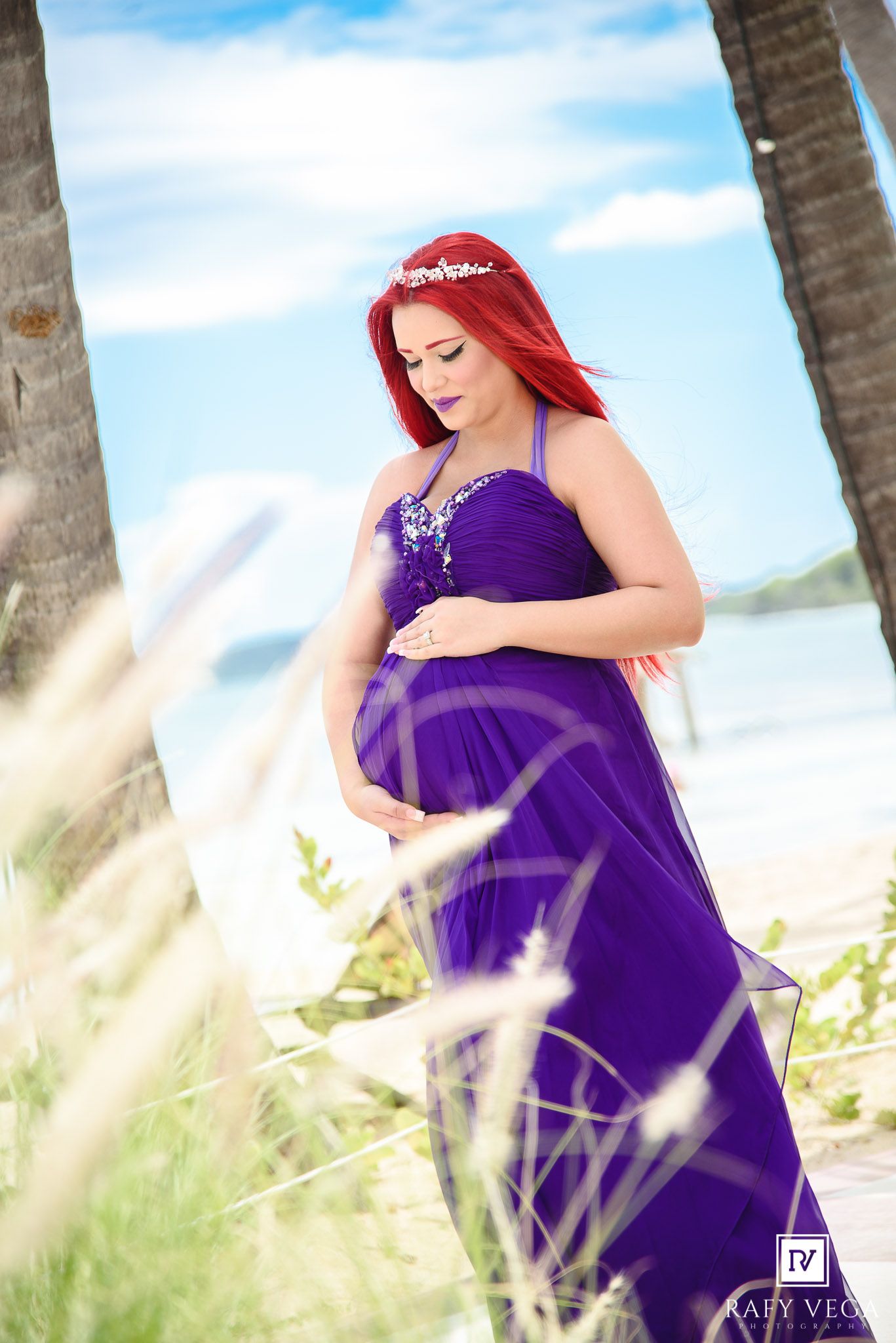 Pregnant woman in purple gown with red hair holding baby bump on beach.