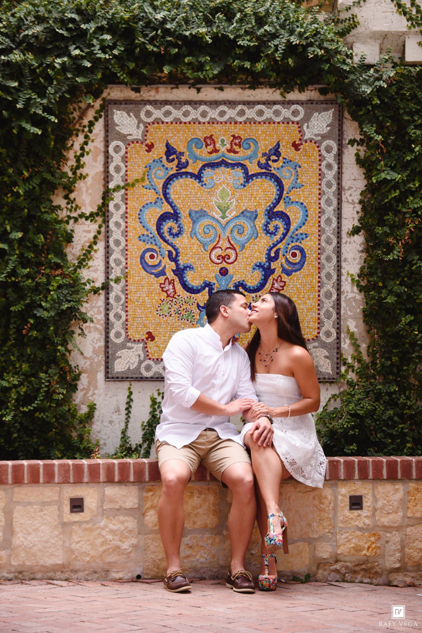 Couple sharing a romantic kiss on a bench in front of a colorful mosaic tile wall surrounded by lush greenery.