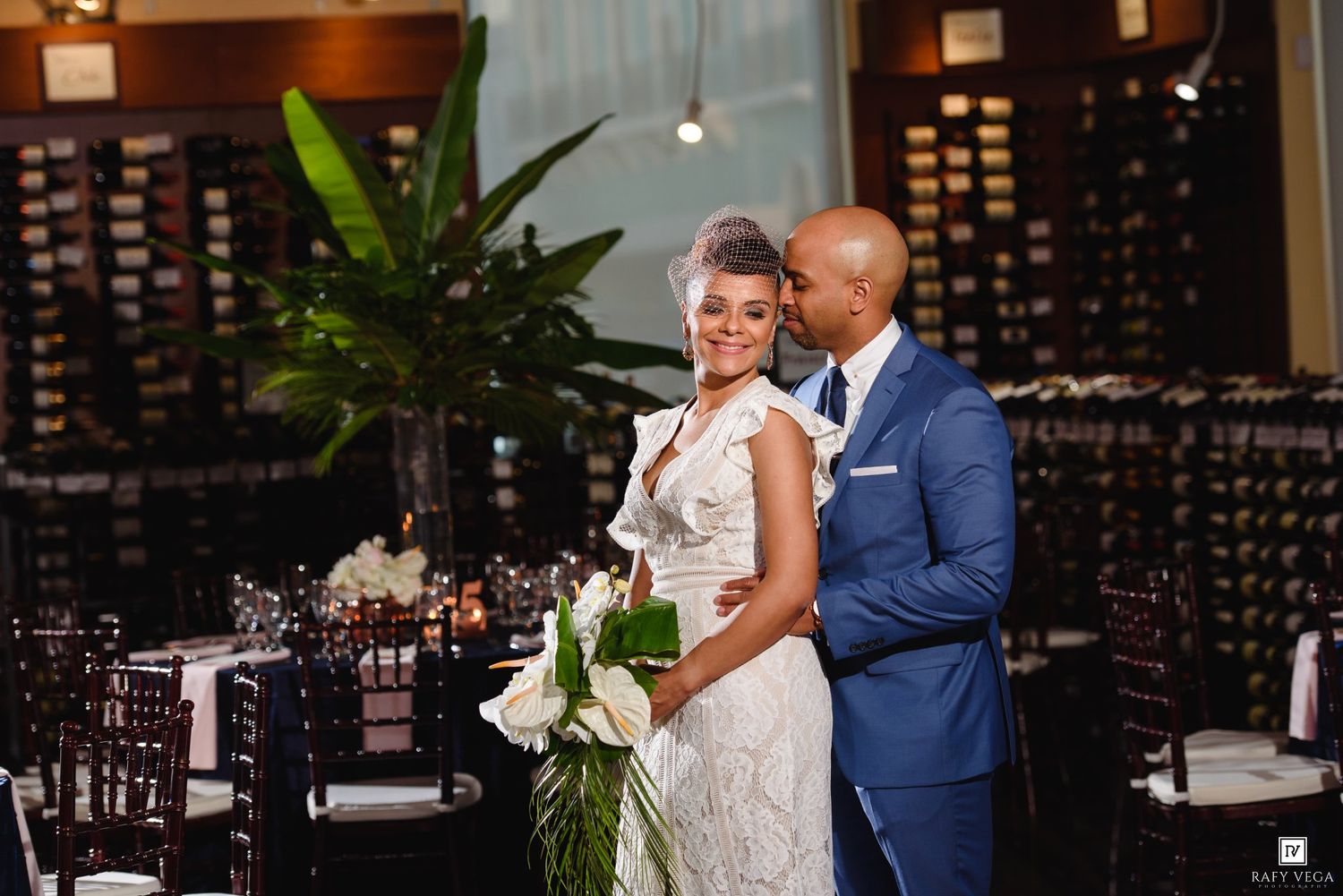 Elegant wedding couple poses in a sophisticated restaurant venue with wine walls, the bride holding white flowers.