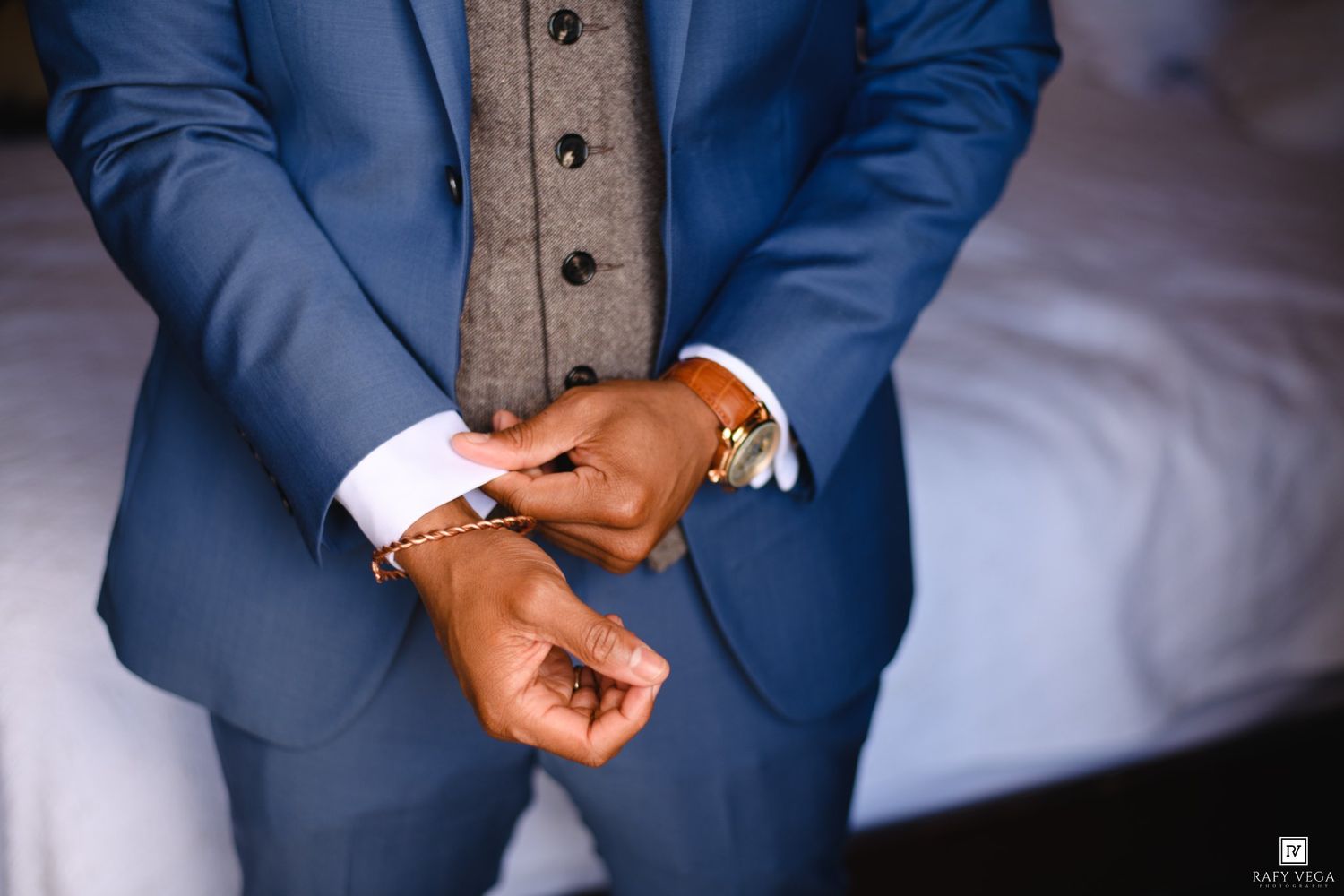 Man in blue suit adjusting cufflinks, wearing a watch and bracelet, with a grey vest underneath.