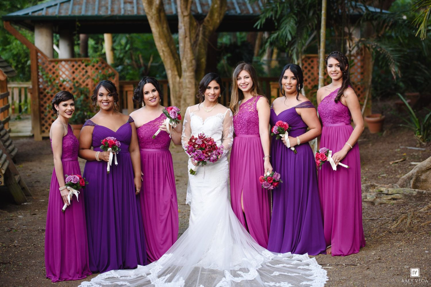Bride in lace gown stands with six bridesmaids wearing purple ombre dresses in a garden setting.
