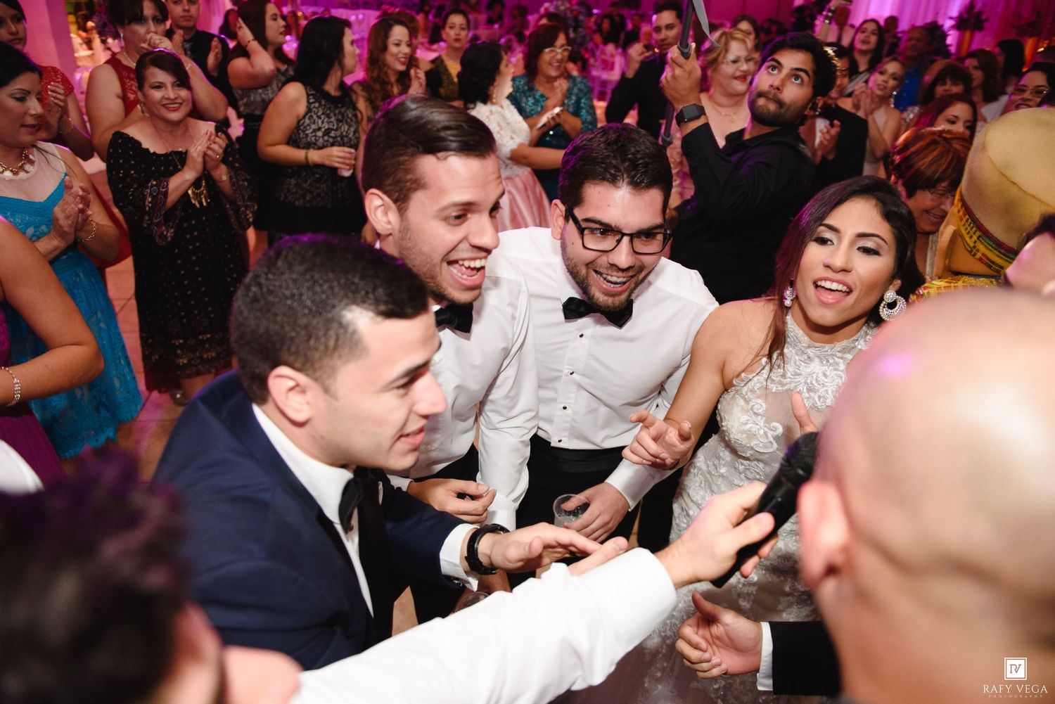 Joyful wedding reception guests dancing and celebrating together in elegant formal attire under pink ambient lighting.