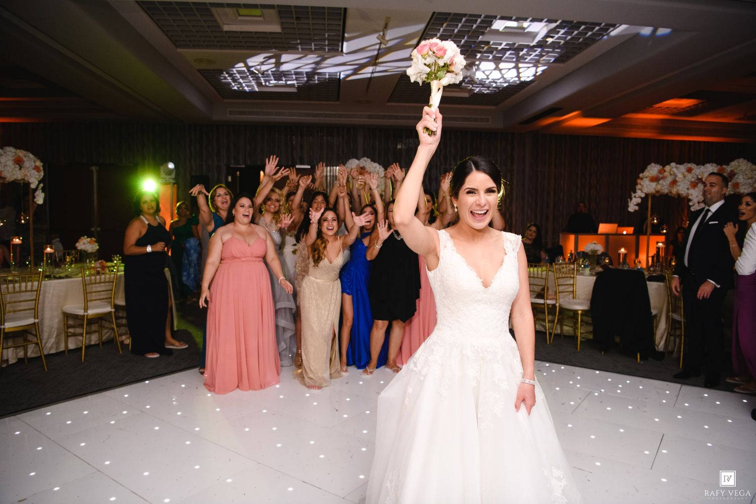 Bride raising bouquet on white dance floor before bouquet toss at elegant wedding reception.