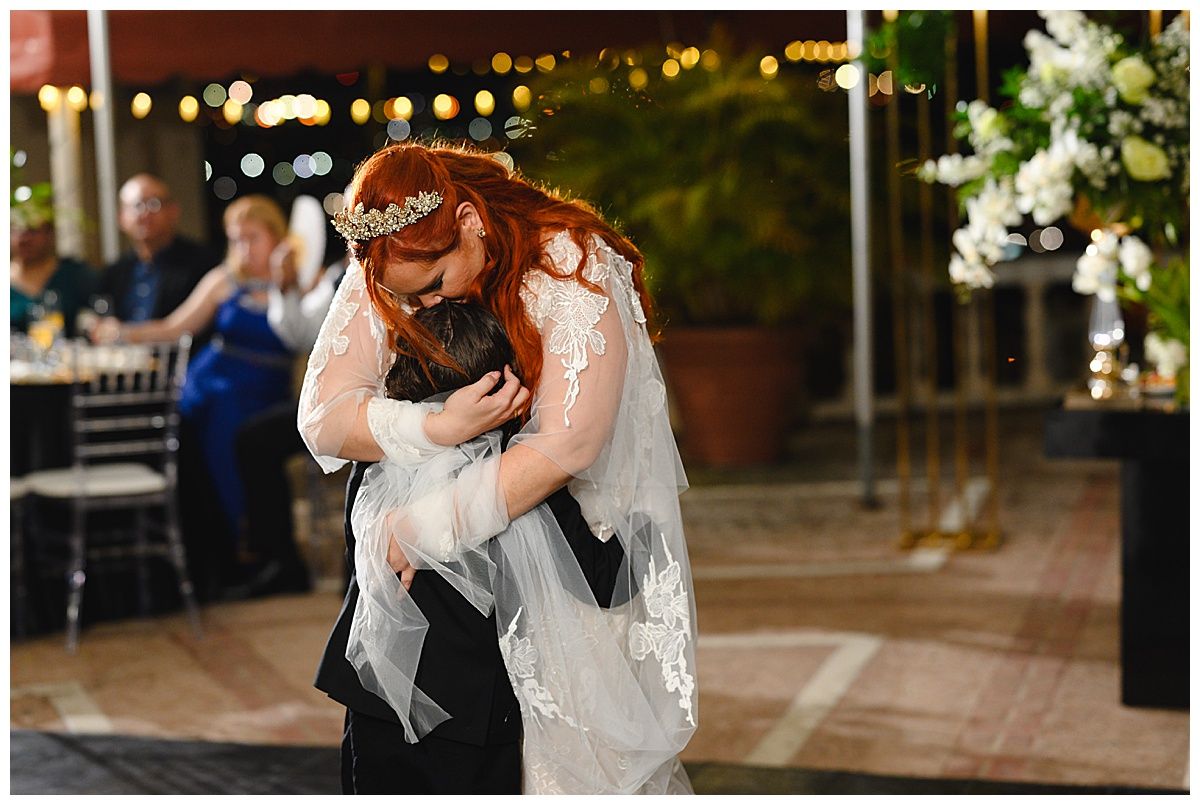 Bride in lace gown and tiara shares emotional first dance hug with groom at wedding reception.