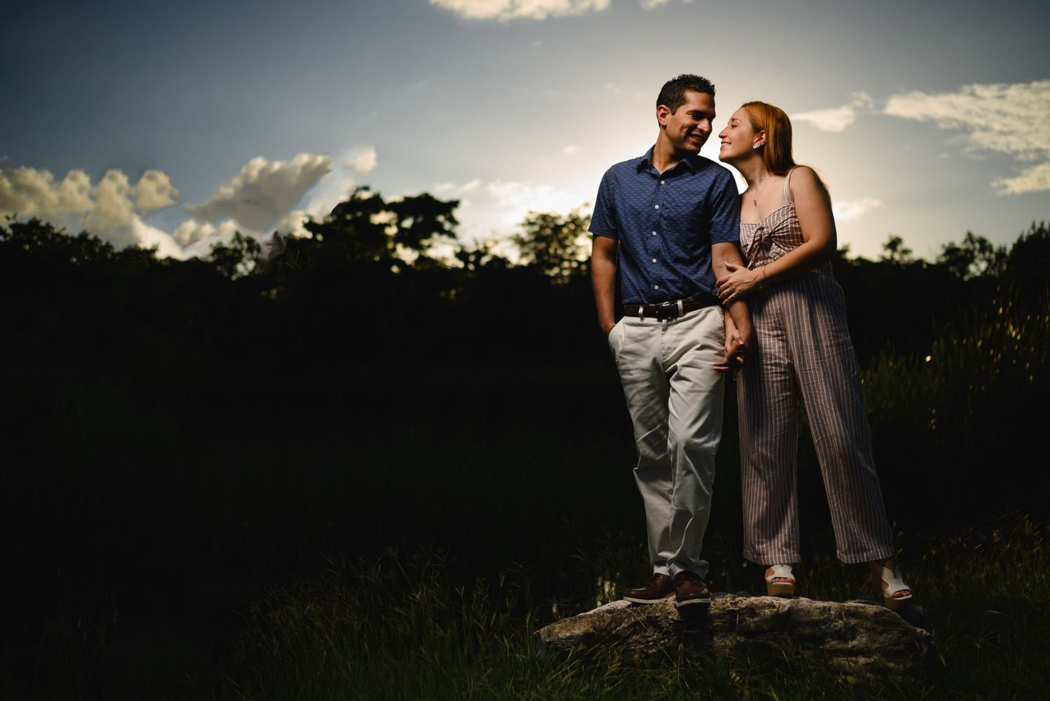 Couple standing together on a rock at sunset with dramatic sky and lush trees in the background.
