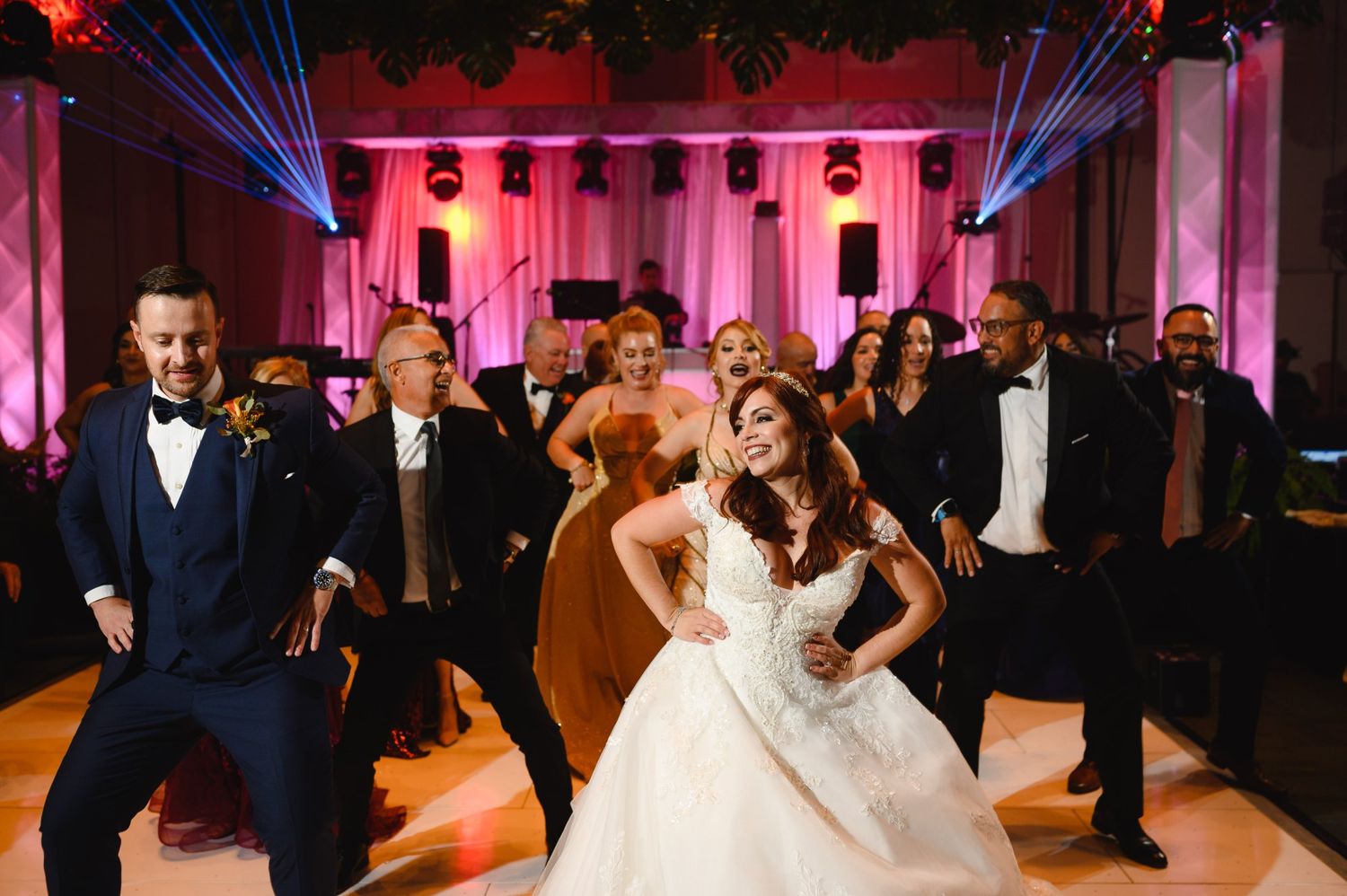 Bride and groom celebrate with wedding party in lively dance floor moment under pink reception lighting.