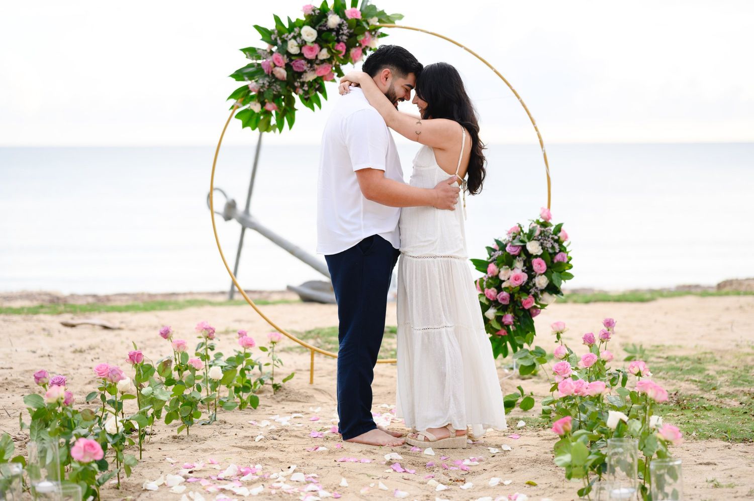Couple embracing under floral hoop arch on beach, surrounded by pink roses, romantic outdoor wedding ceremony.