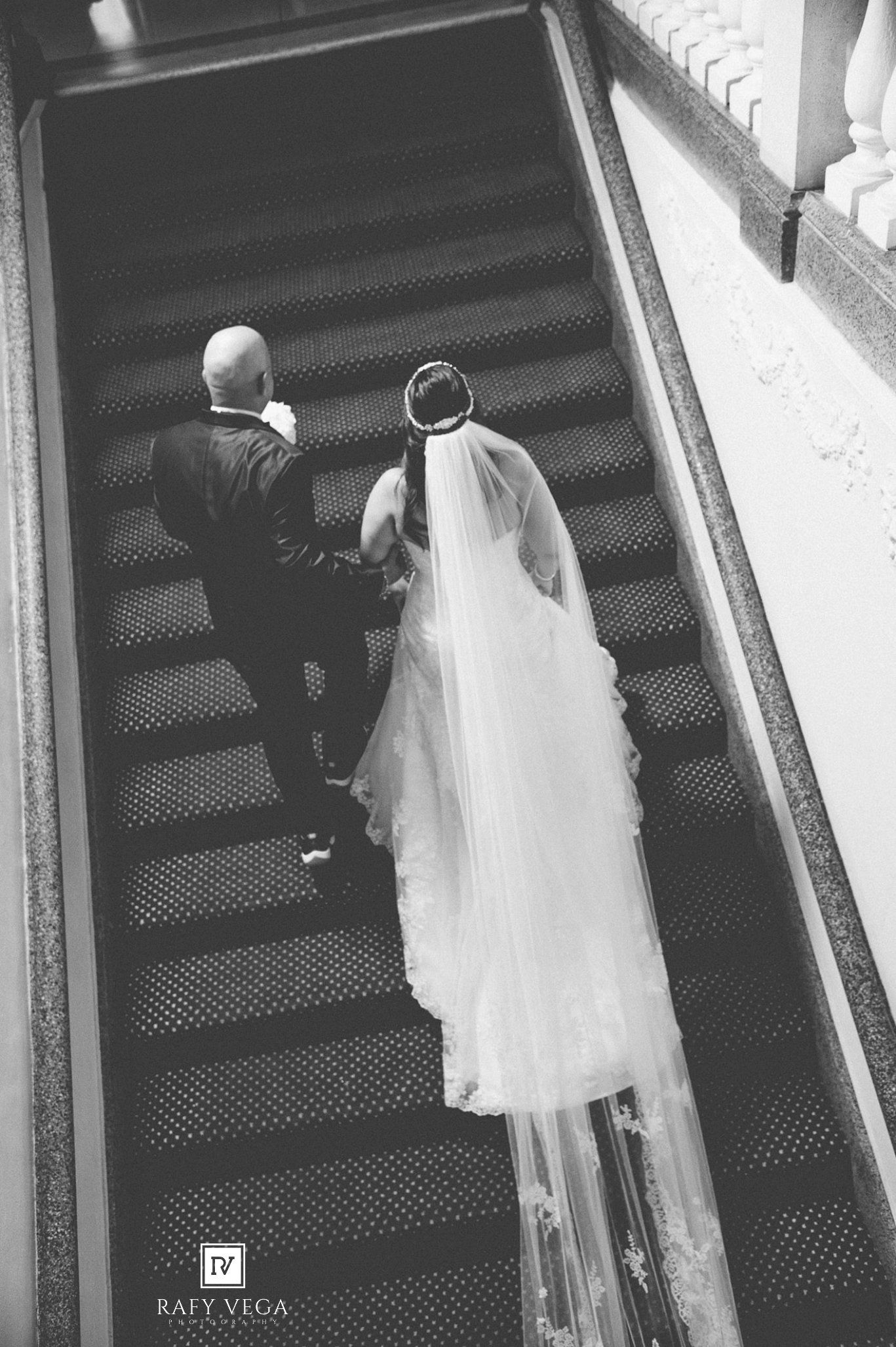 Aerial black and white photo of bride in flowing veil and groom walking down elegant staircase at wedding.