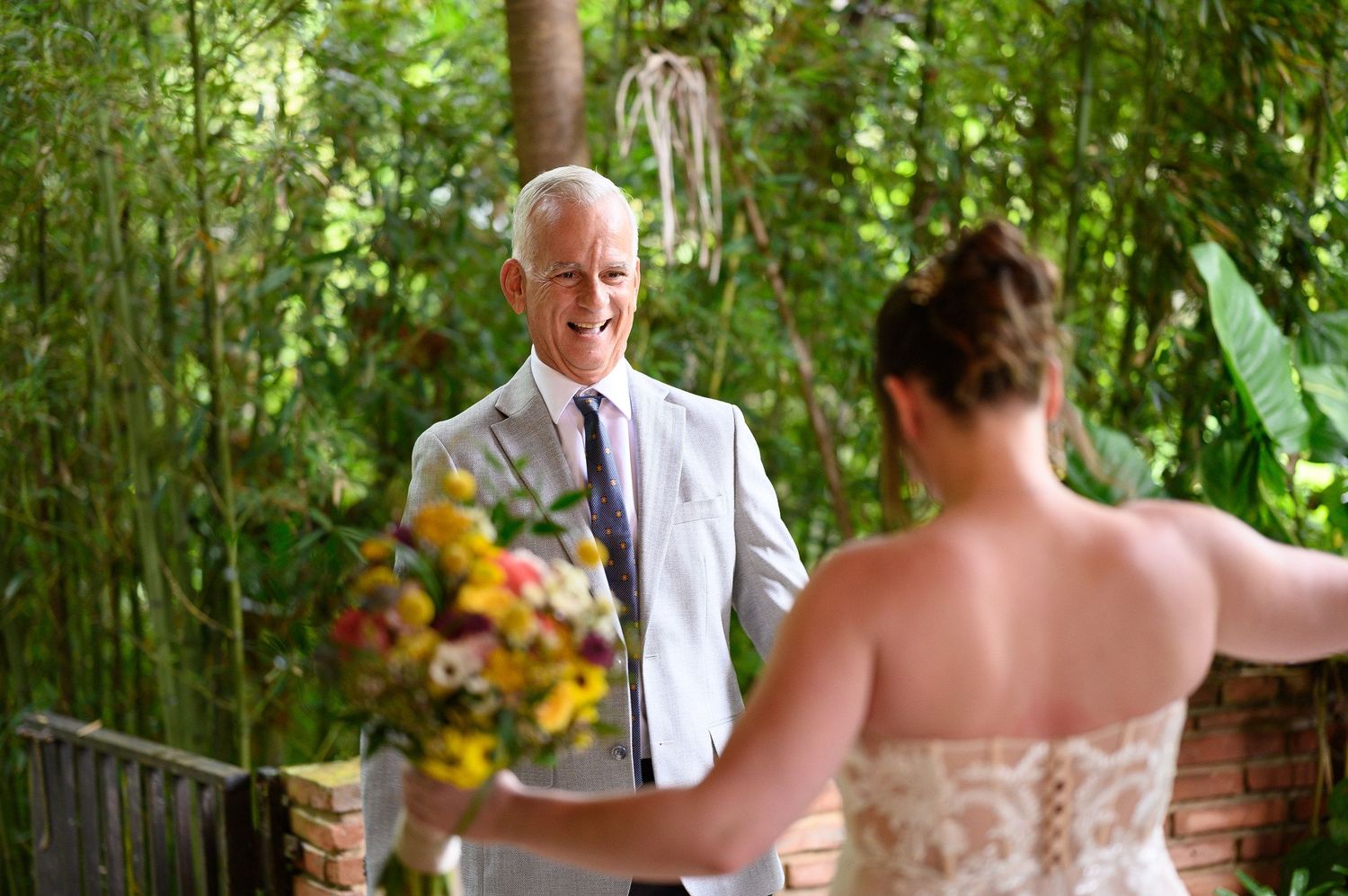 Groom in white suit smiles at bride holding colorful bouquet during outdoor wedding ceremony surrounded by lush greenery.