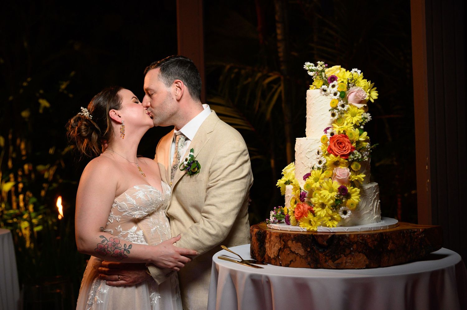 Bride and groom share a kiss beside a tall floral wedding cake on a rustic wood stand at their reception.