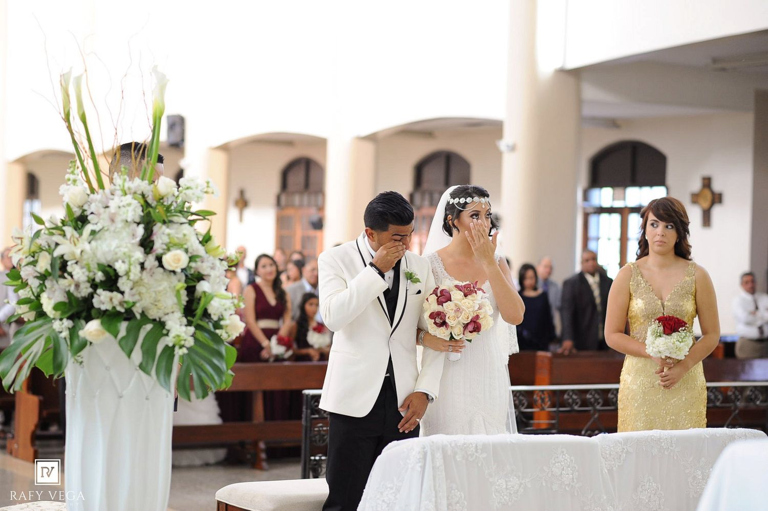 Groom kisses bride's hand at church altar during wedding ceremony, bridesmaid holding bouquet nearby.