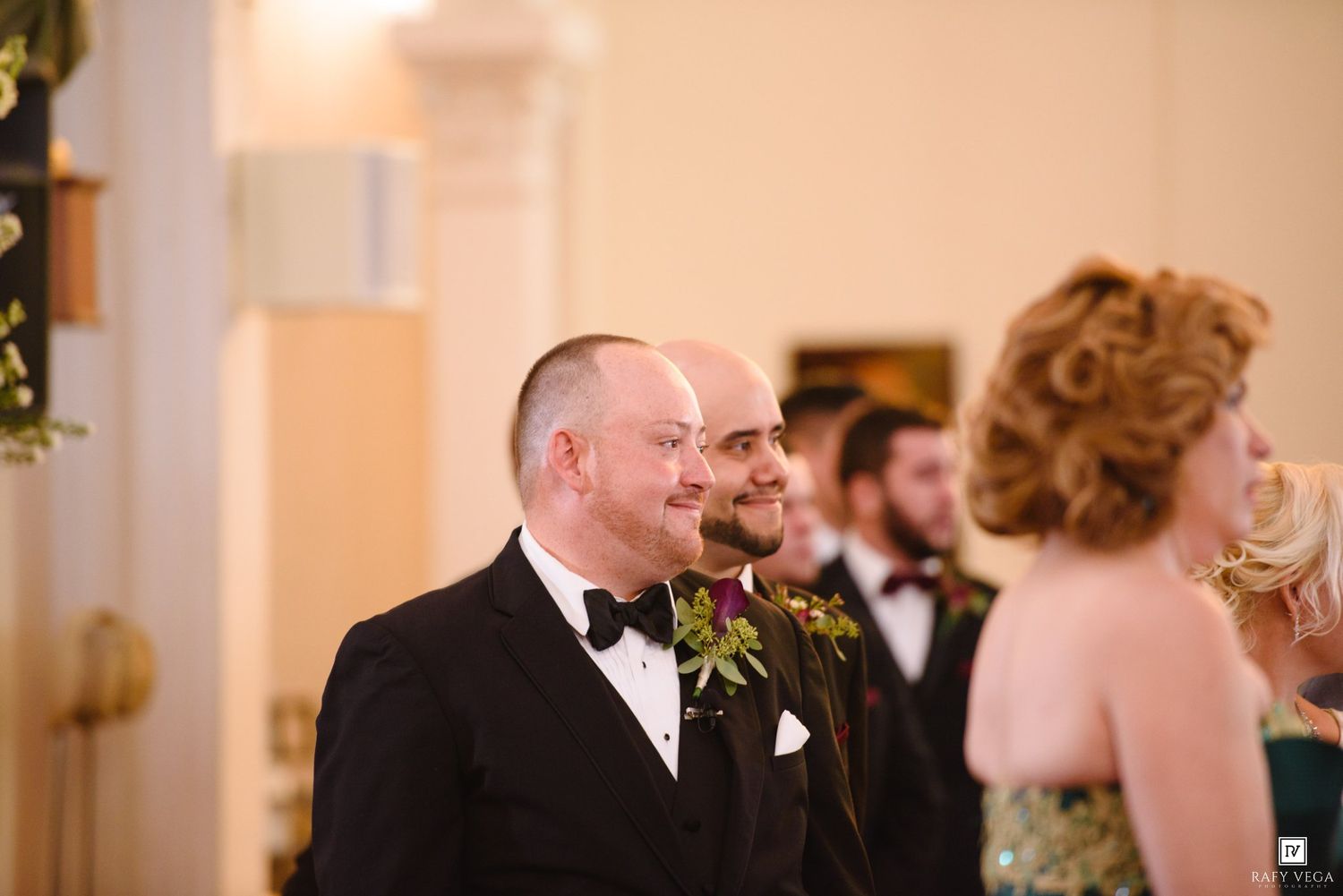 Groom in black tuxedo smiling at wedding ceremony with groomsmen and guests in elegant venue.