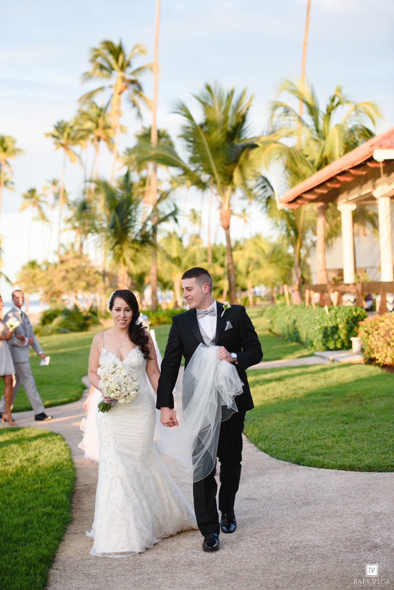 Bride and groom walk hand-in-hand on a tropical resort path, surrounded by palm trees, as groom holds her veil.