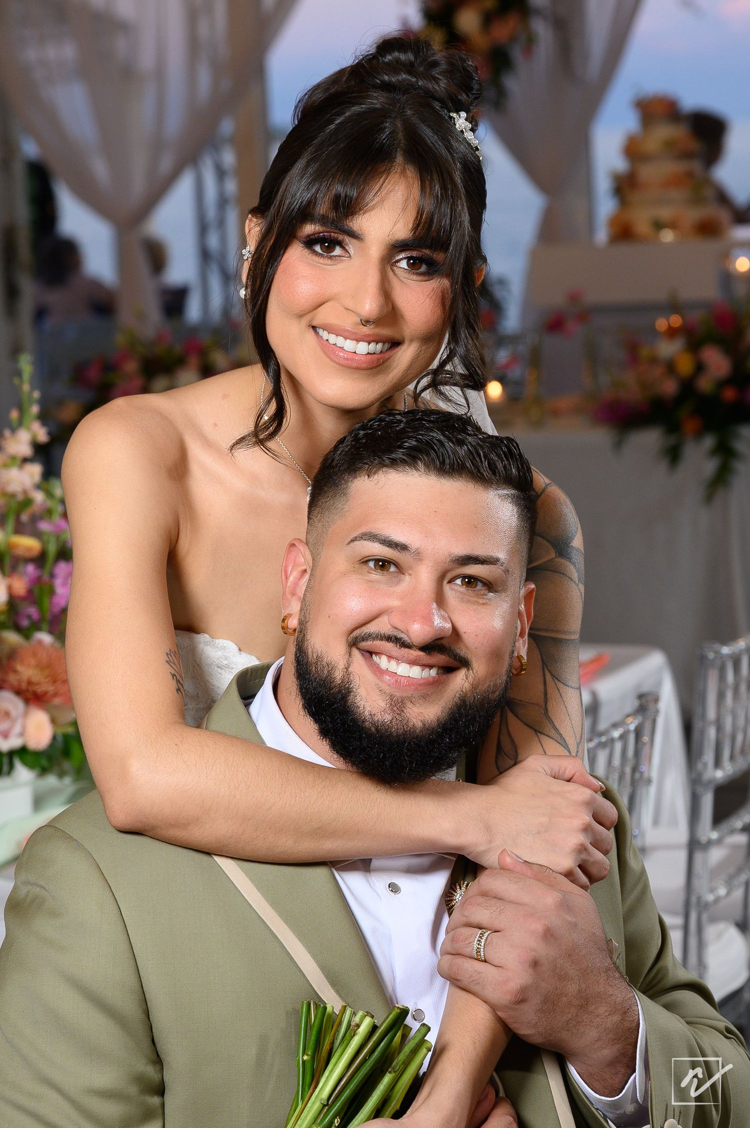 Smiling bride in a strapless dress hugs her bearded groom in a tan suit from behind at a wedding reception with floral decor.