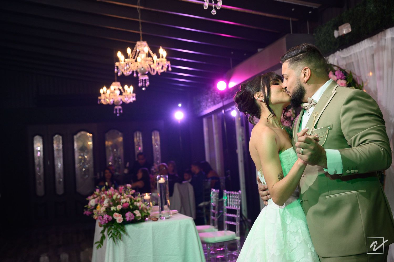 Bride in a light green gown and groom in an olive suit share a romantic kiss at a wedding reception with chandeliers and a floral table.