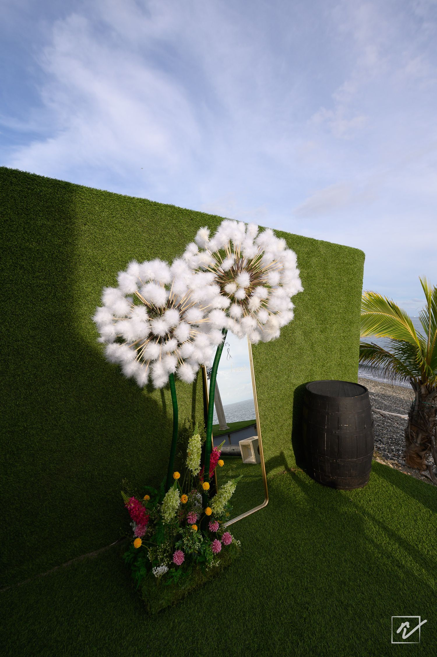 Giant white dandelion flower sculptures and a colorful floral arrangement beside a mirror on artificial grass near a barrel and palm tree.