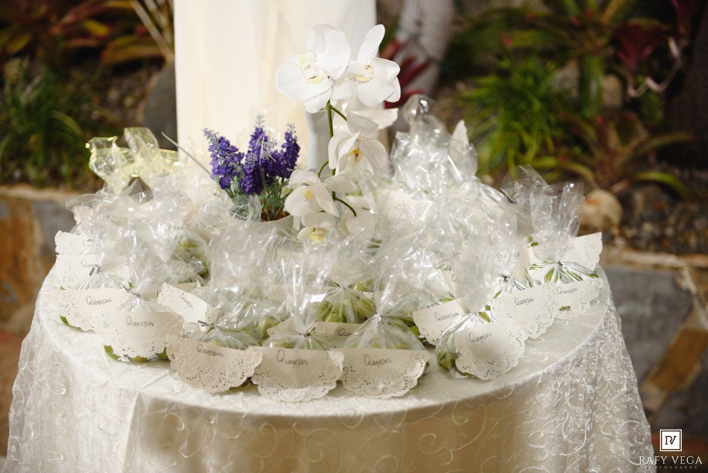 White wedding favor table with wrapped gifts, orchids, and purple hyacinth flowers on lace tablecloth.