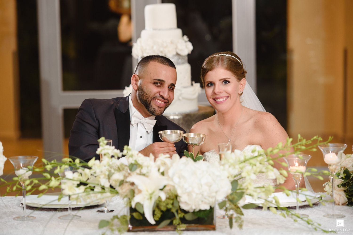 Bride and groom smiling at sweetheart table with white floral centerpieces and wedding cake in background.