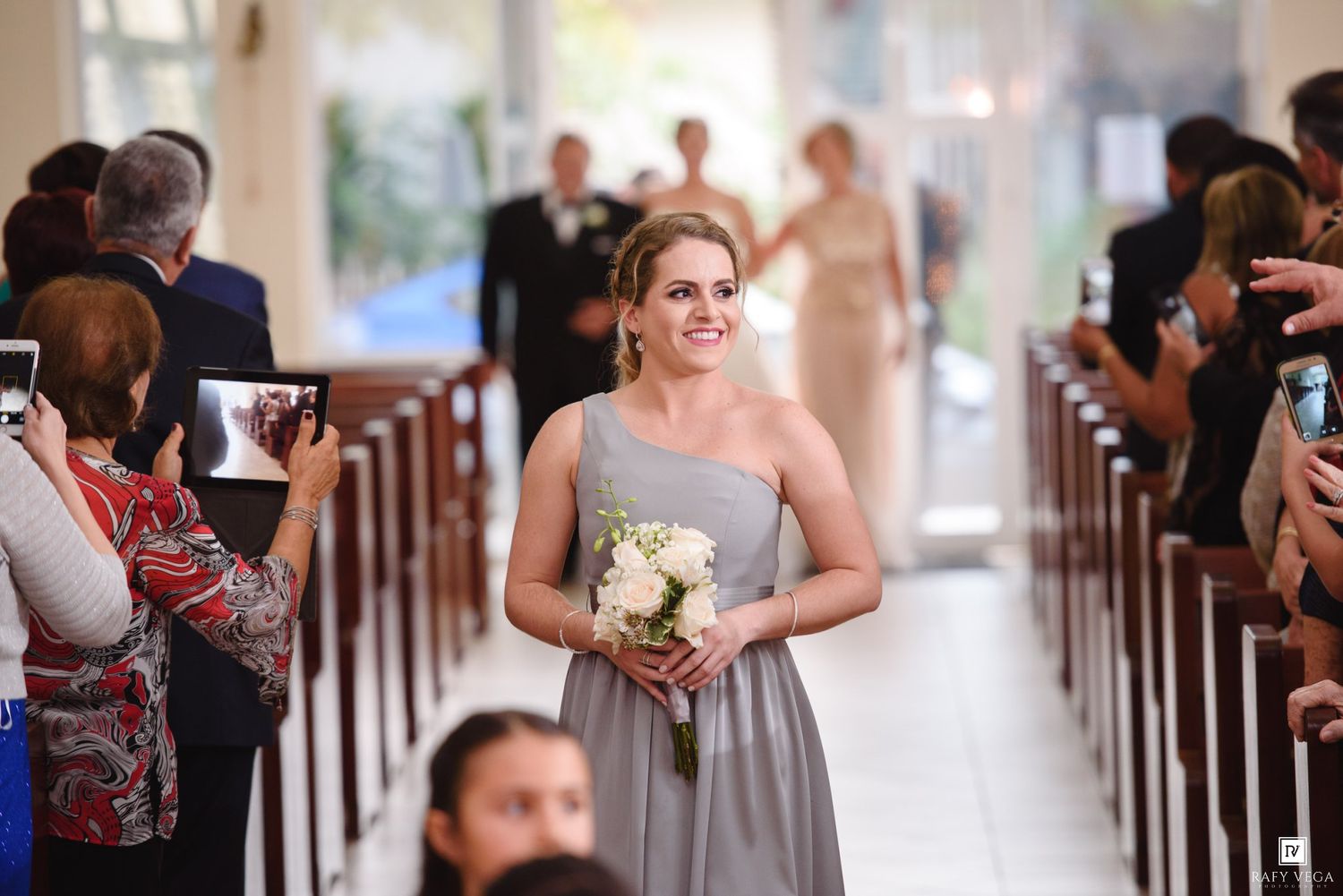 Smiling bridesmaid in gray dress walks down church aisle holding white bouquet during wedding ceremony.