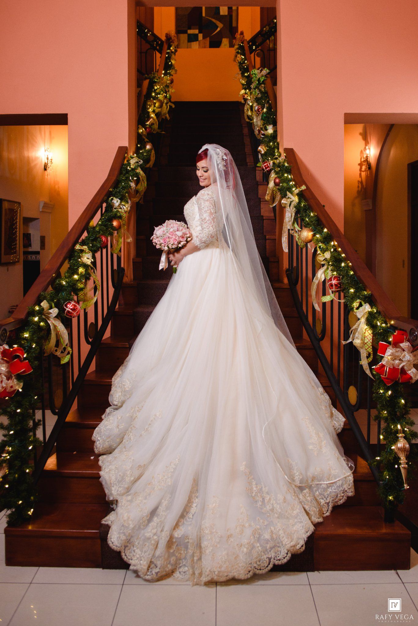 Bride in elegant lace ball gown with veil poses on Christmas-decorated staircase holding pink bouquet.