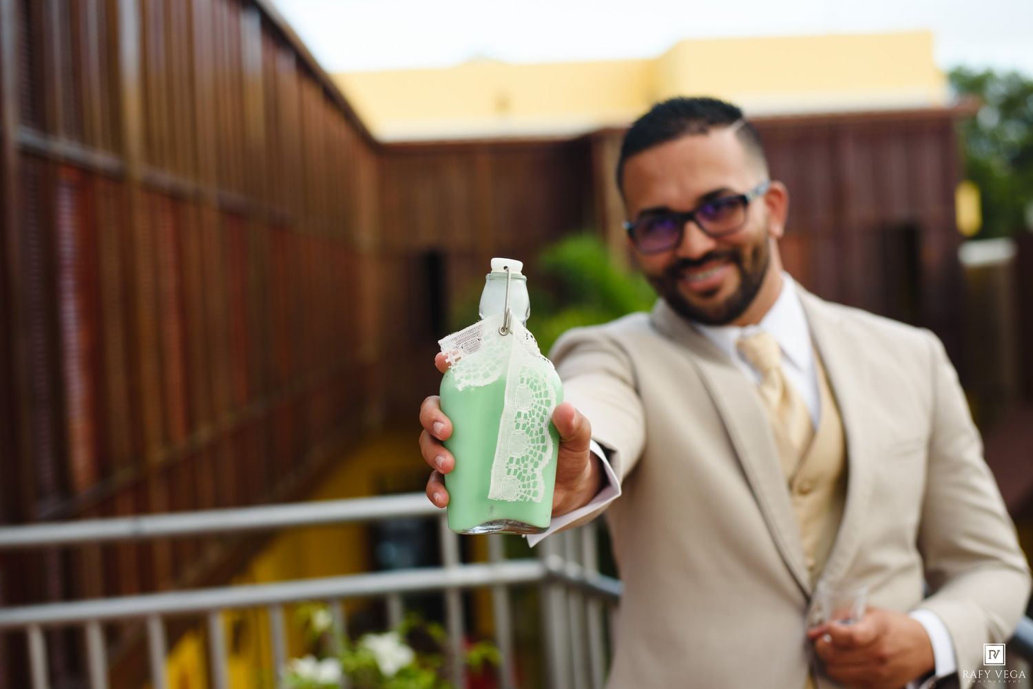 Smiling man in beige suit holding a green bottle outdoors on a balcony.