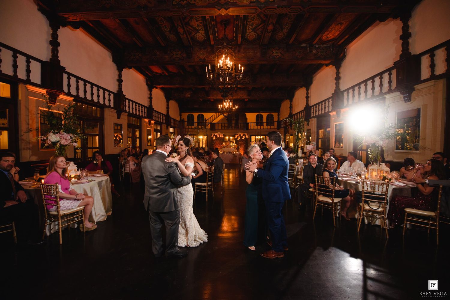 Couples share first dance in elegant ballroom with chandeliers and guests seated at decorated tables during wedding reception.