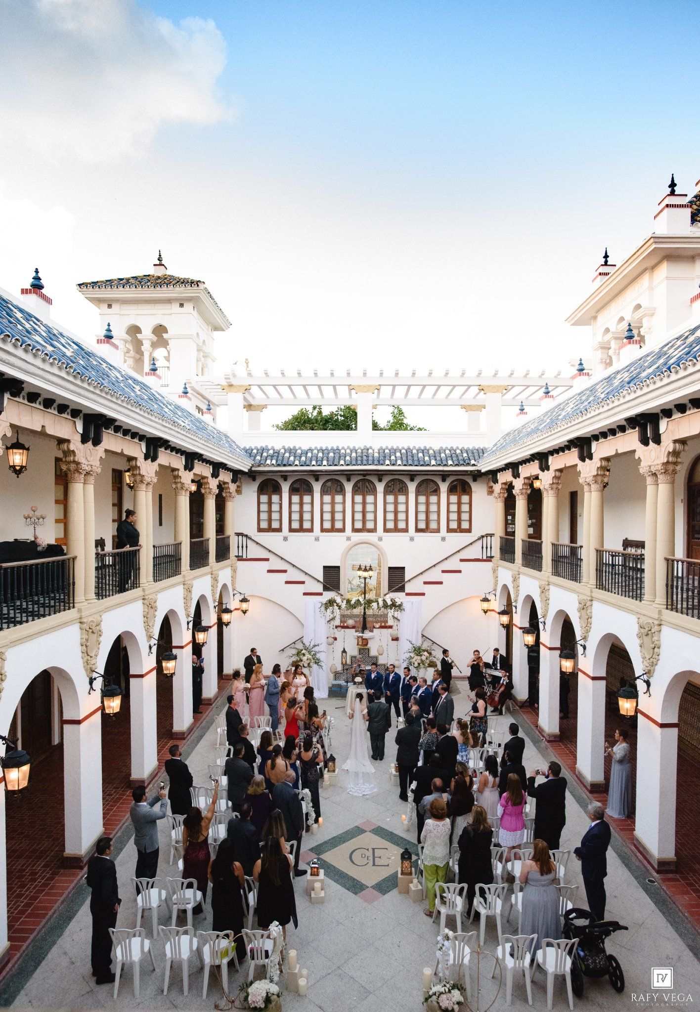 Elegant outdoor courtyard wedding ceremony at a Spanish-style venue with arched walkways and white chairs.