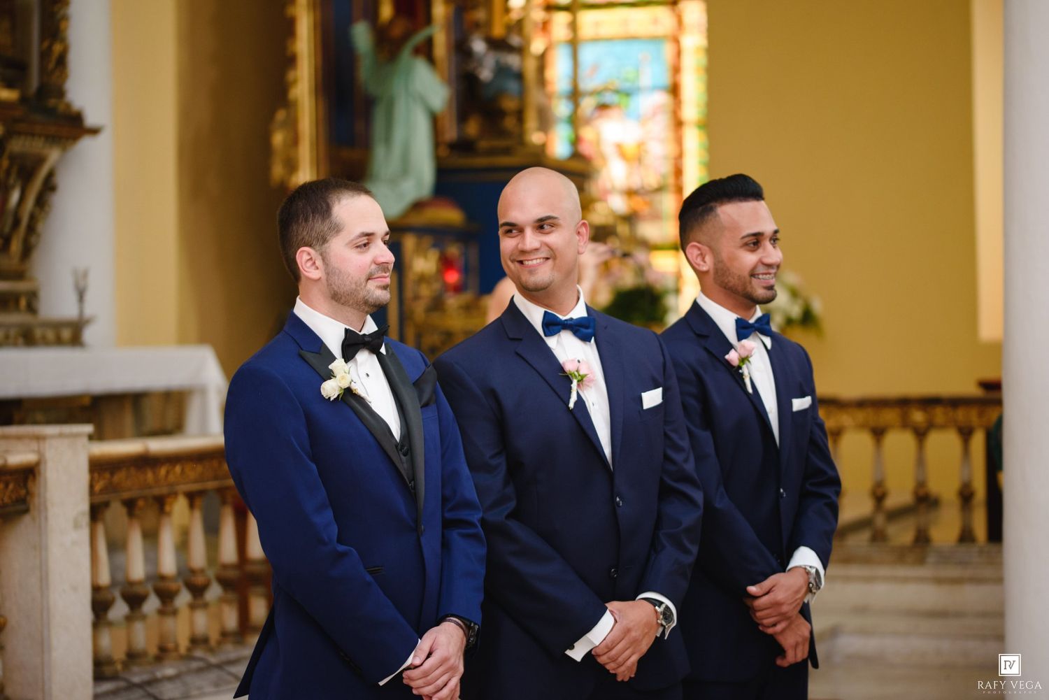 Three groomsmen in matching navy blue tuxedos standing at church altar before wedding ceremony.