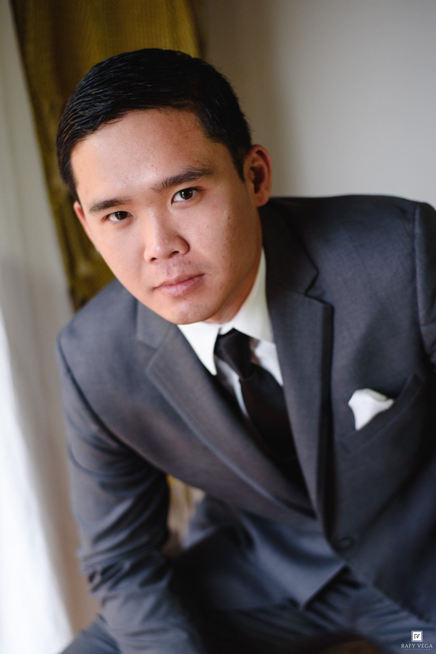 A young Asian groom in a sharp gray suit with white pocket square and black tie poses confidently for a wedding portrait.