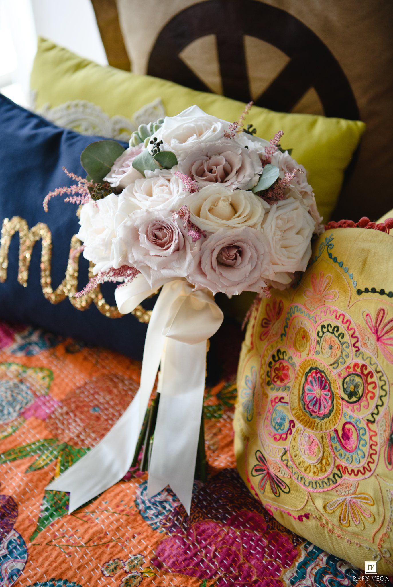 A romantic bridal bouquet of blush and mauve roses with eucalyptus, tied with a white ribbon, resting on colorful embroidered pillows.