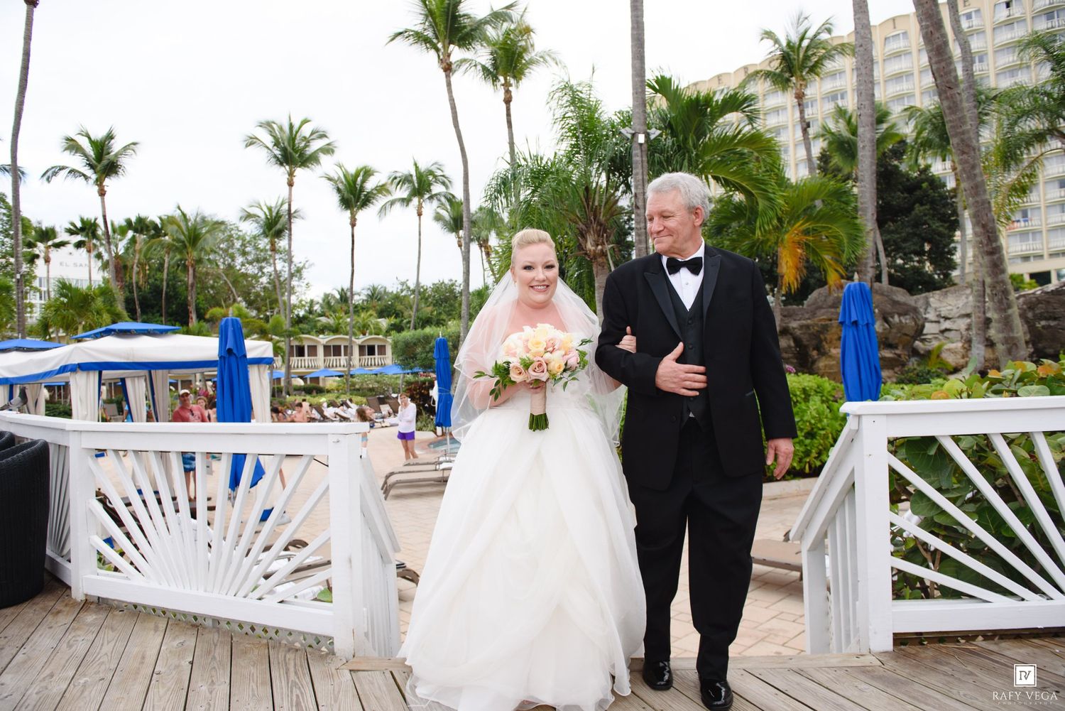 Bride in white gown walks with older man in tuxedo on wooden dock at tropical resort wedding.